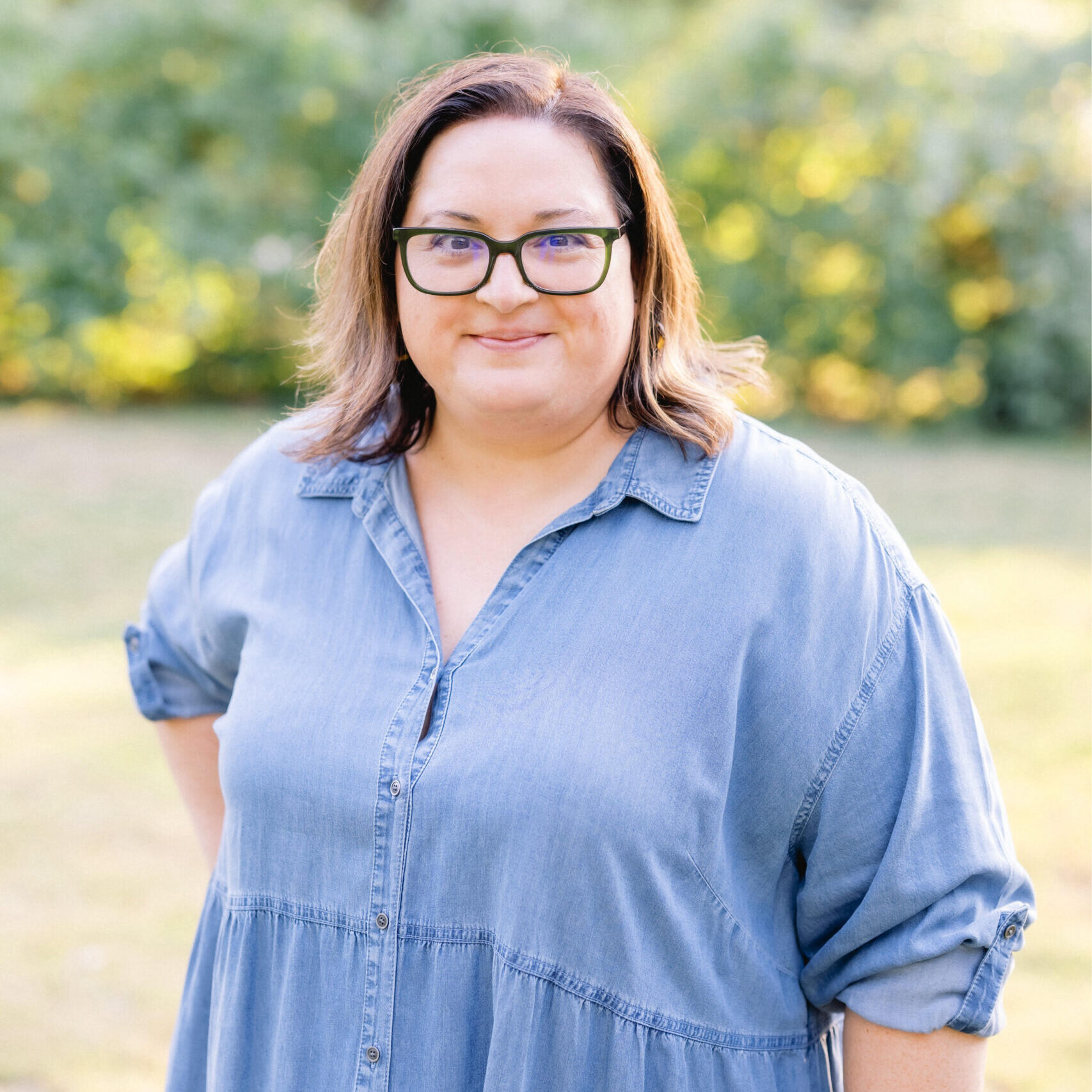 A professional head shot of a woman with dark hair, wearing glasses and a blue dress smiling at the camera
